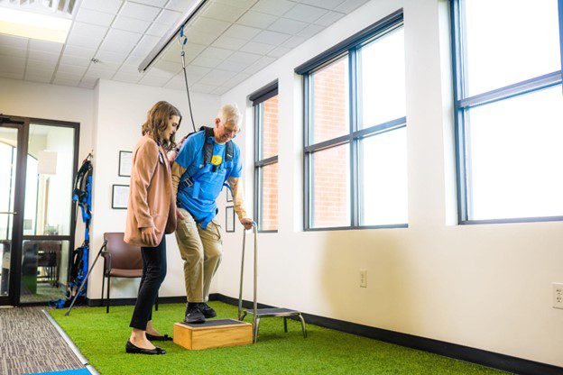 A man in a solo-step harness steps onto a box with the help of a physical therapist