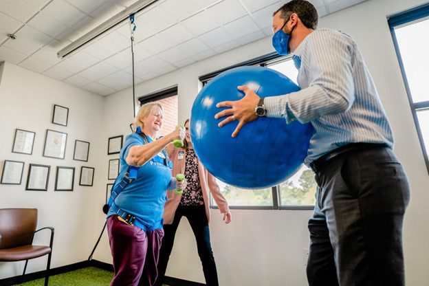 A patient in a solo-step harness is using hand weights, and a therapist holds an exercise ball