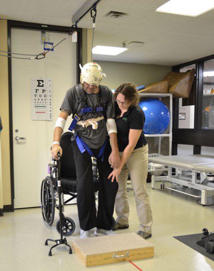 A physical therapist helps a spinal patient stand up from their wheelchair, while using an overhead harness for safety